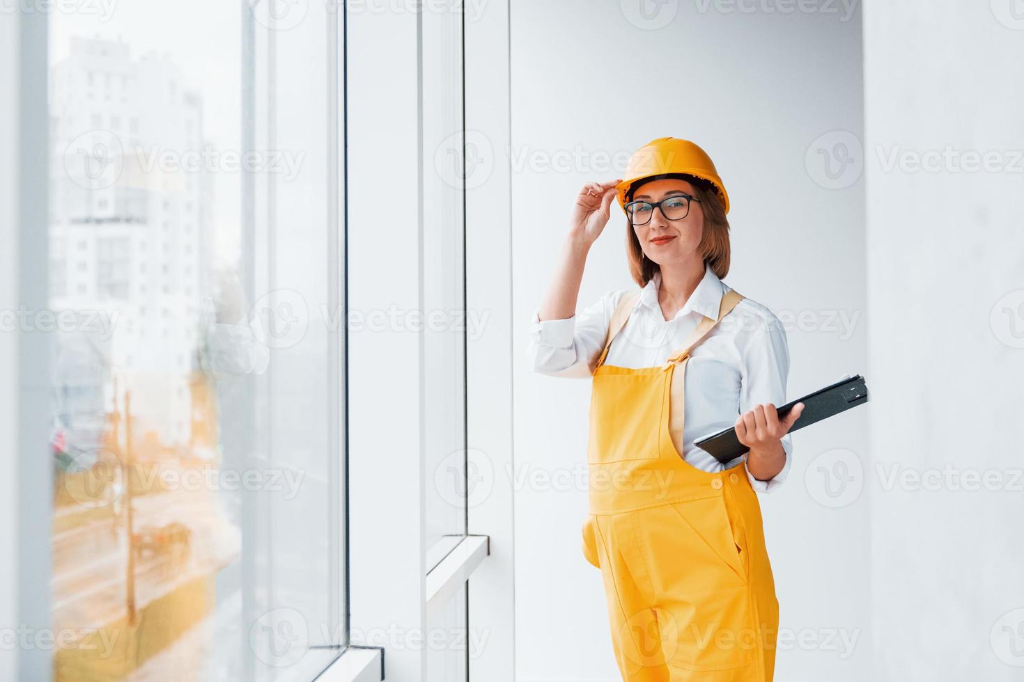 With notepad in hands. Female worker or engineer in yellow uniform and hard hat standing indoors photo