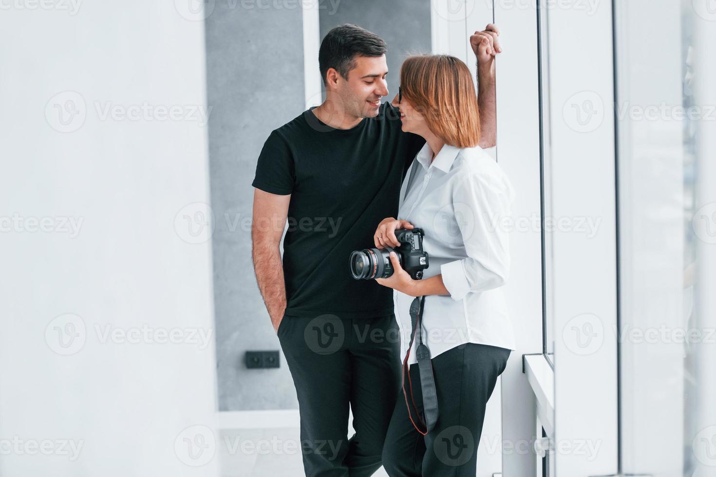 Woman in formal clothes and with camera in hand standing inside of empty room with man photo