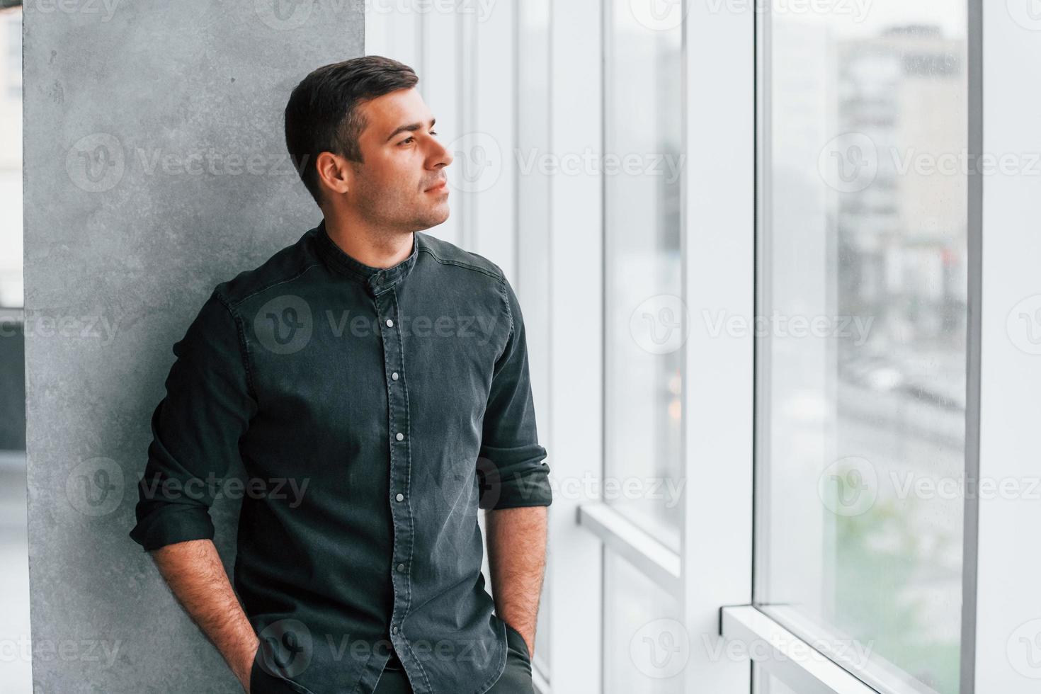 Elegant man in black shirt leaning on the wall indoors and looking through the big window photo