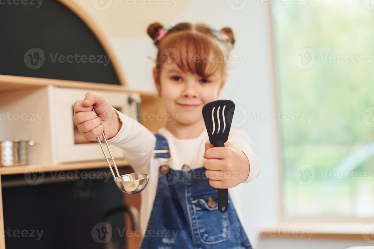 Holding tools in hands and shows it. Little girl in casual clothes have fun by playing with toys on the kitchen photo