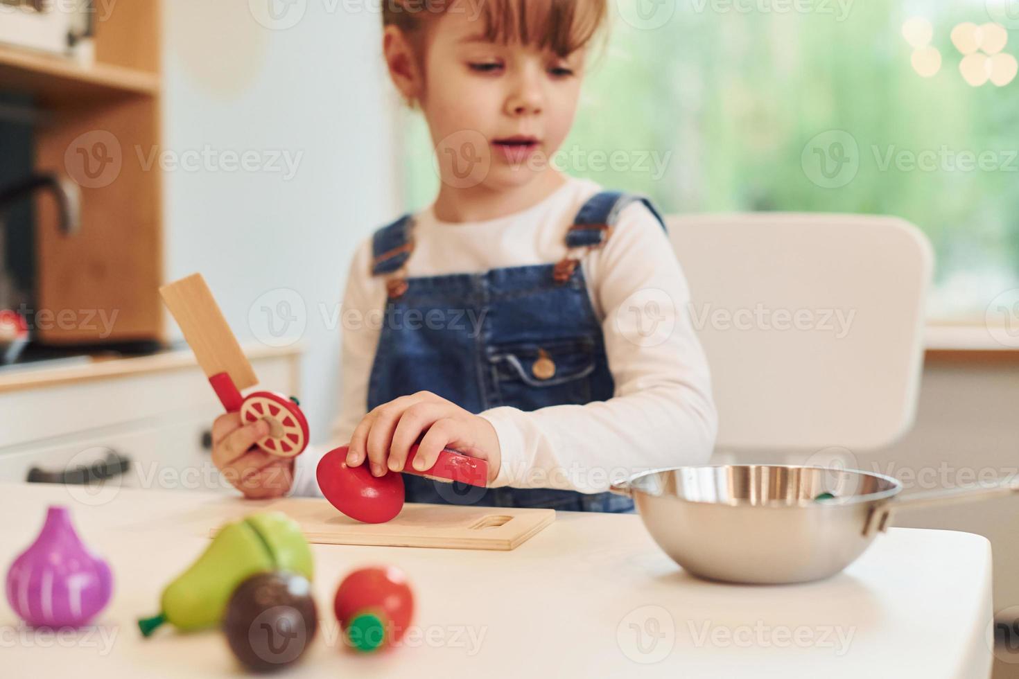 Little girl in casual clothes sits by table and having fun by playing with toys on the kitchen photo