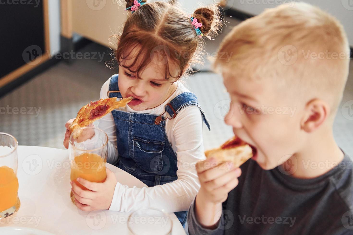 tres niños sentados en el interior junto a la mesa y comiendo pizza con jugo de naranja juntos foto