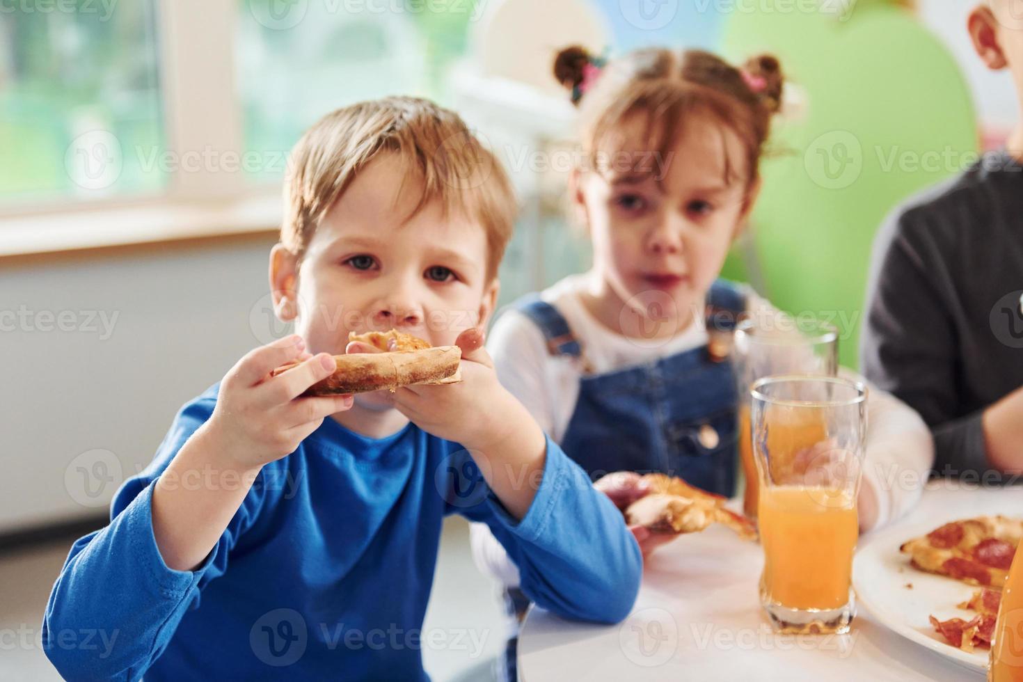 Three children sitting indoors by the table and eating pizza with orange juice together photo