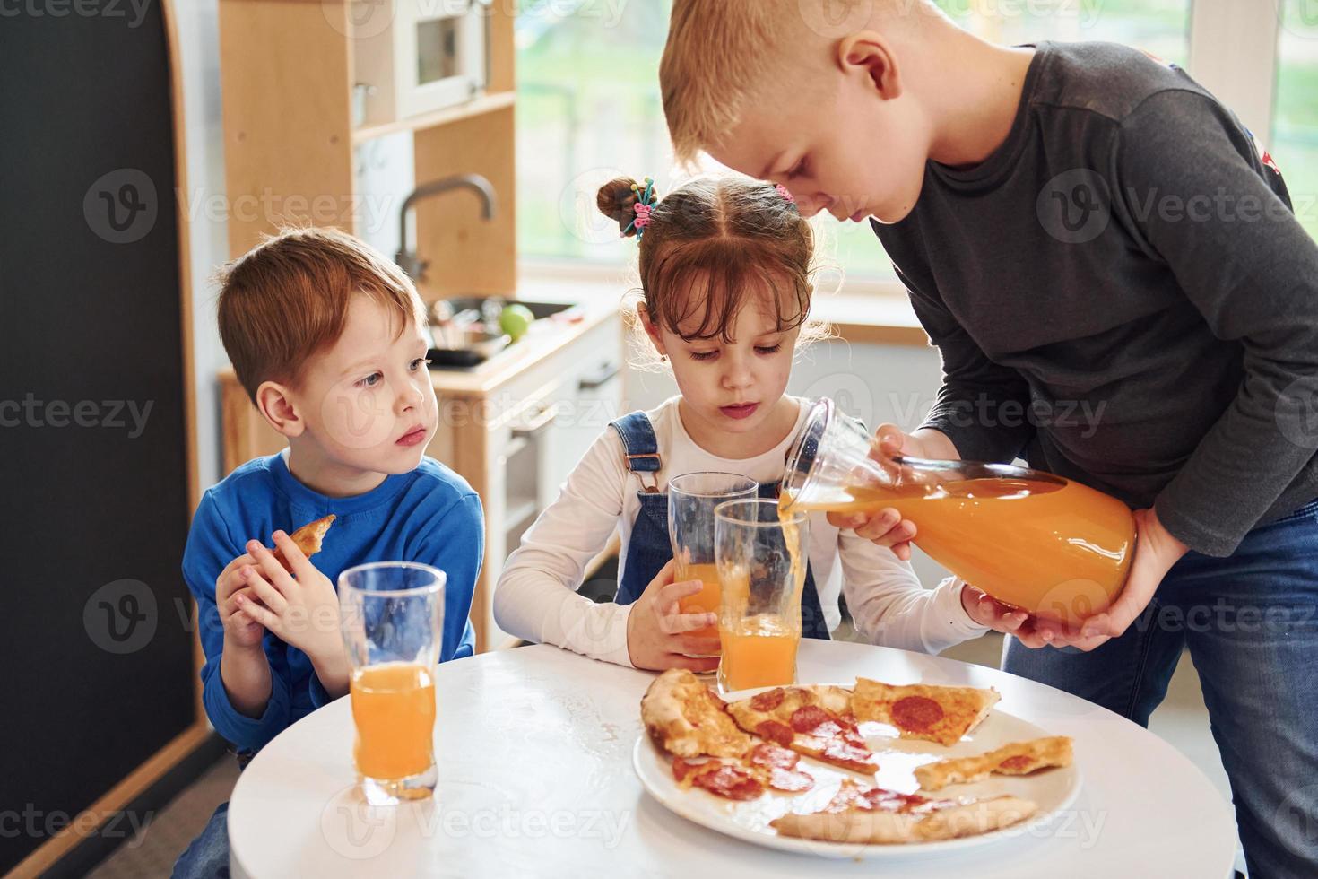 tres niños sentados en el interior junto a la mesa y comiendo pizza con jugo de naranja juntos foto