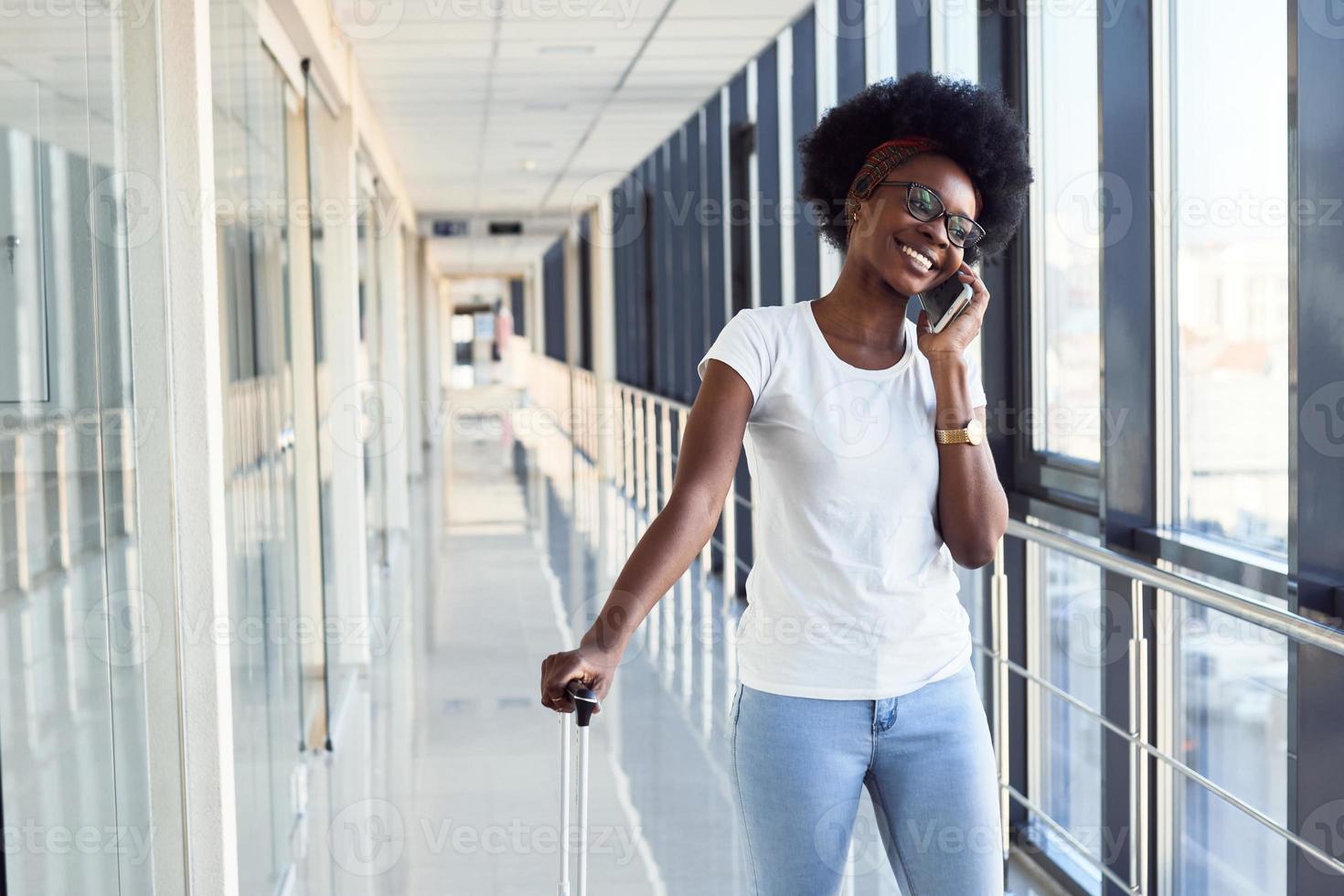 una joven pasajera afroamericana con ropa informal está en el aeropuerto con teléfono foto