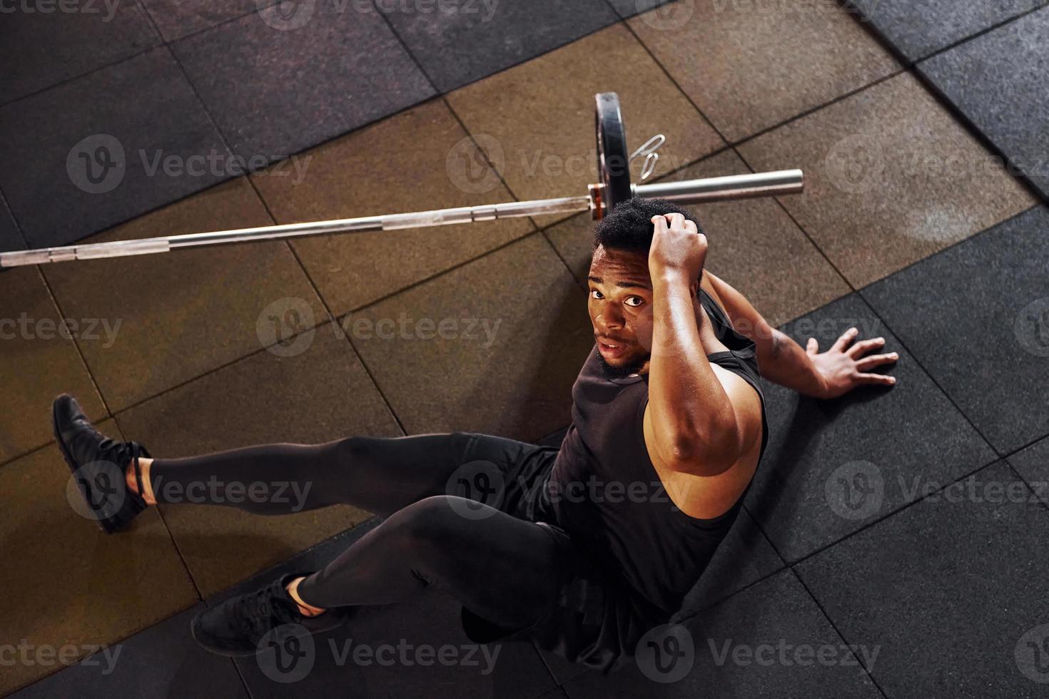 Sitting on the floor. Strong african american man in sportive clothes have workout day in the gym photo