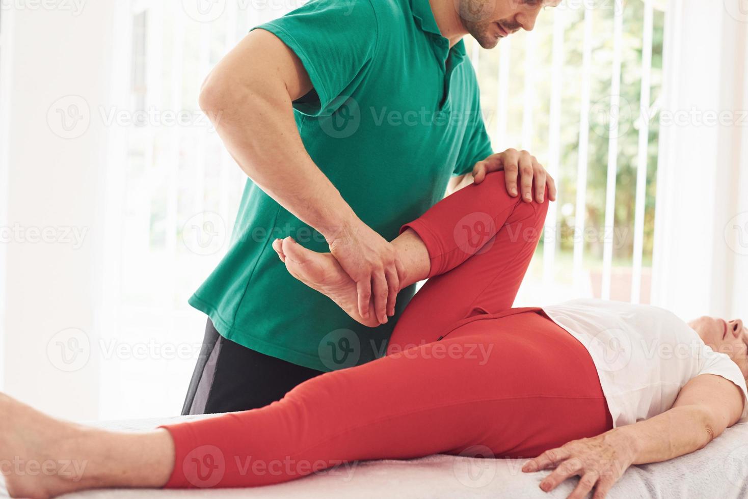 Older woman have a visit in clinic, lying down and having rehabilitation. Male instructor helping her photo