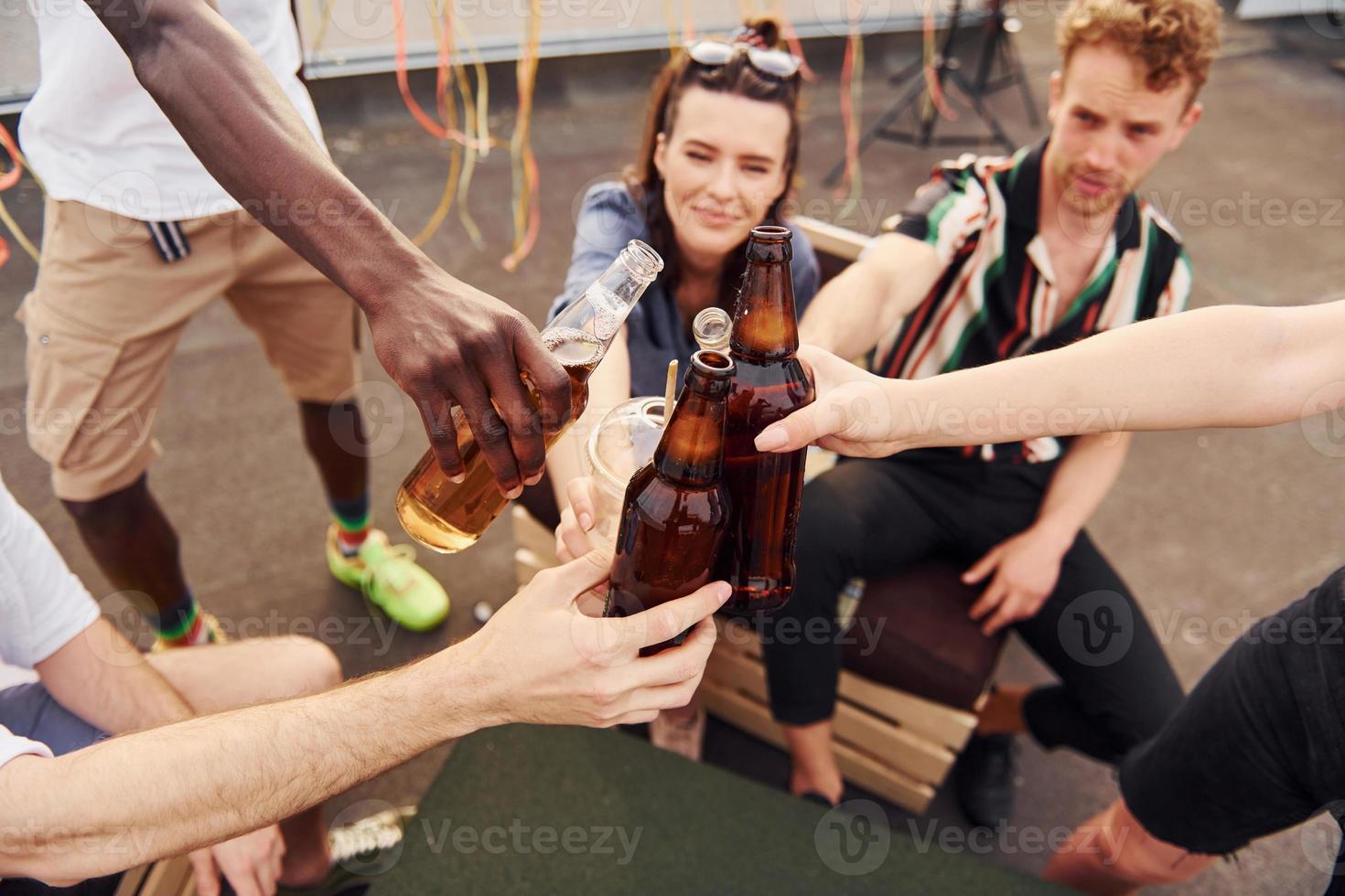 haciendo vítores por botellas con cerveza. un grupo de jóvenes con ropa informal tienen una fiesta en la azotea juntos durante el día foto