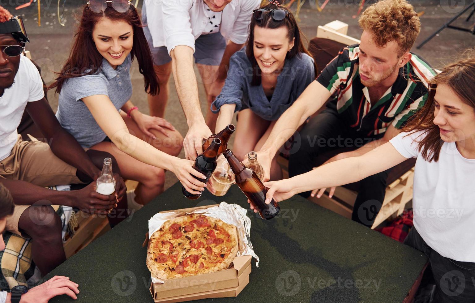 haciendo vítores por botellas con cerveza. un grupo de jóvenes con ropa informal tienen una fiesta en la azotea juntos durante el día foto