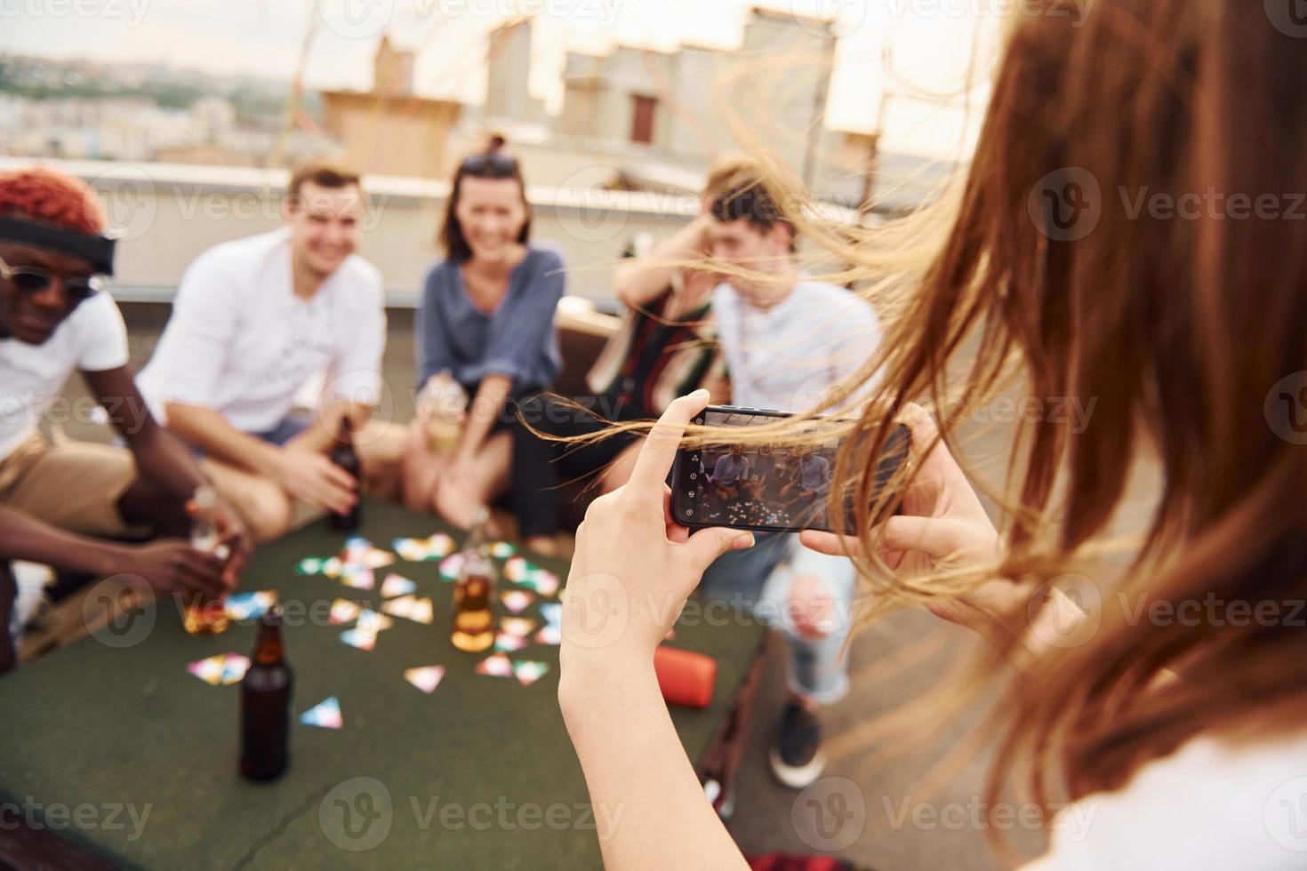 Girl doing photo when people playing card game. Group of young people in casual clothes have a party at rooftop together at daytime