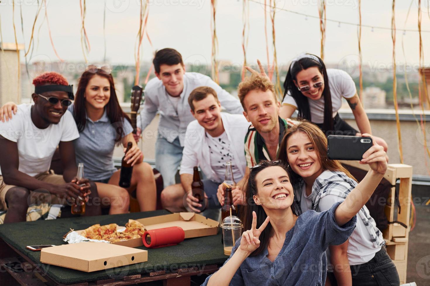 Girl making selfie. With delicious pizza. Group of young people in casual clothes have a party at rooftop together at daytime photo