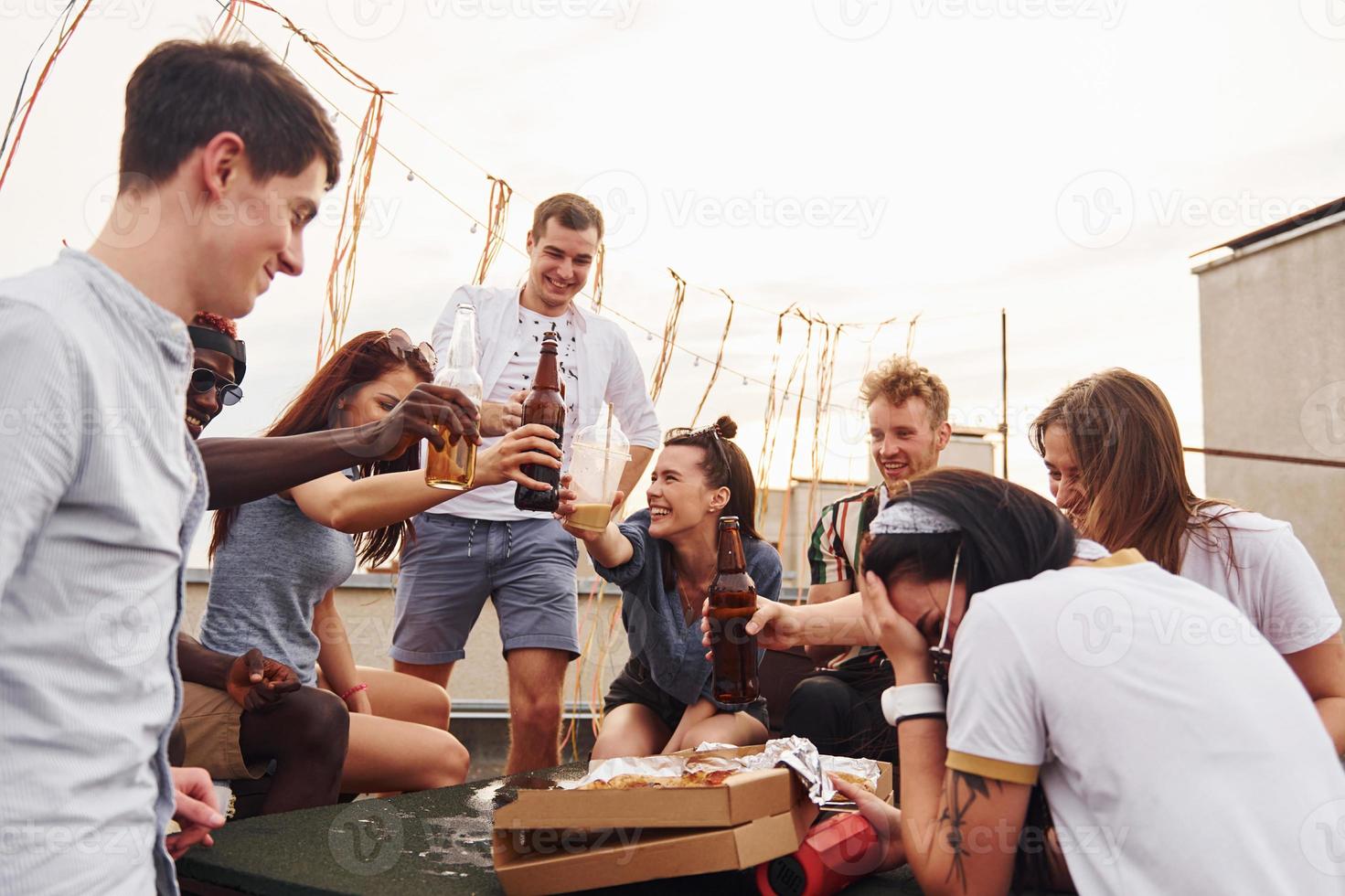 haciendo vítores por botellas con cerveza. un grupo de jóvenes con ropa informal tienen una fiesta en la azotea juntos durante el día foto