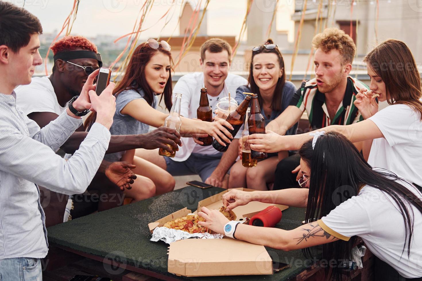 Doing cheers by bottles with beer. Group of young people in casual clothes have a party at rooftop together at daytime photo