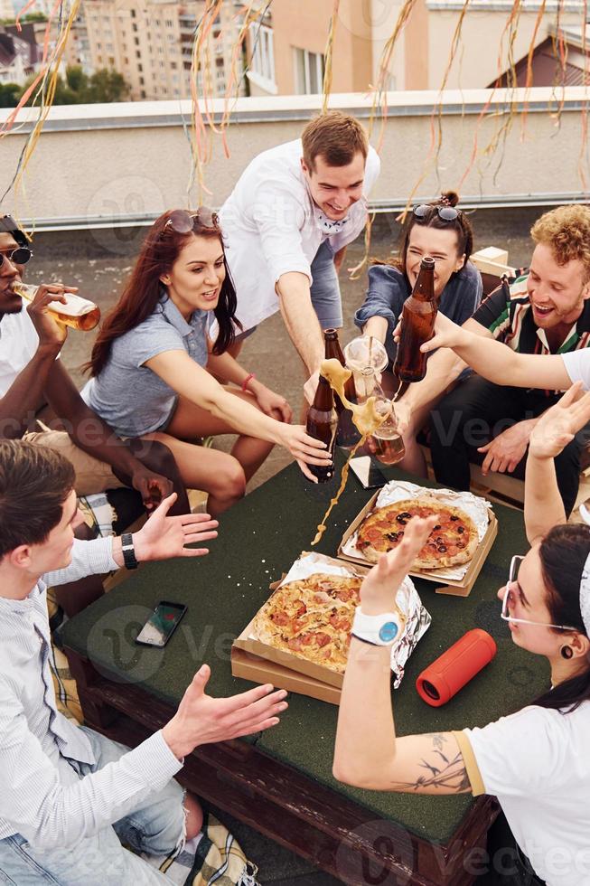 Doing cheers by bottles with beer. Group of young people in casual clothes have a party at rooftop together at daytime photo