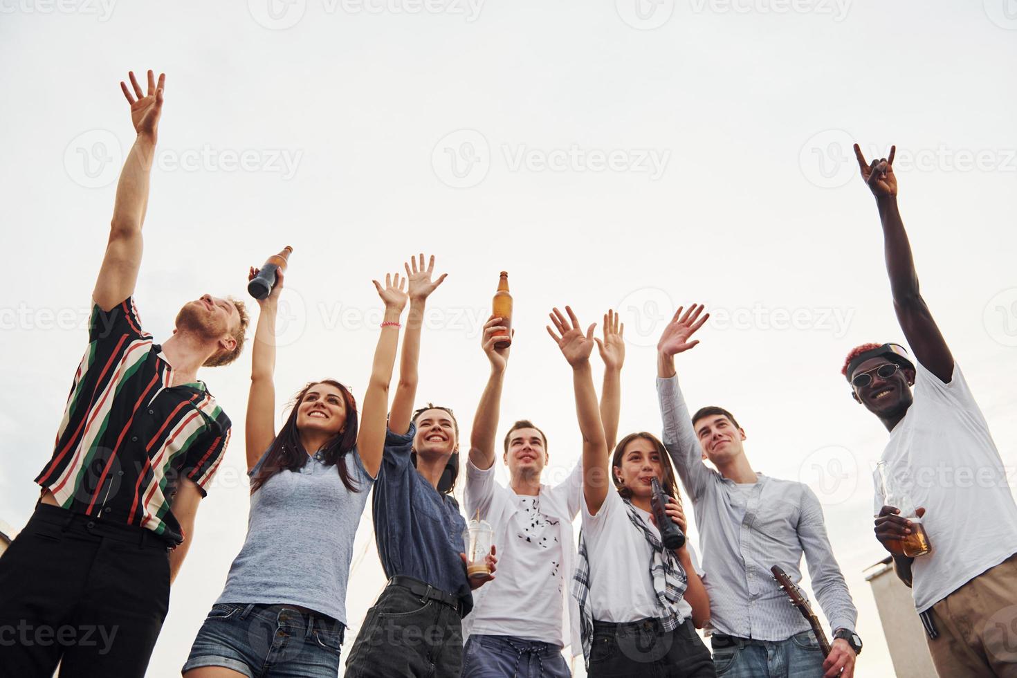 de pie alto y mirando el cielo nublado. un grupo de jóvenes con ropa informal tienen una fiesta en la azotea juntos durante el día foto