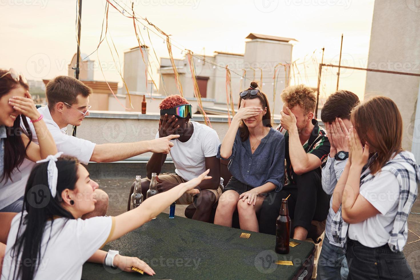 Sitting by table and playing card game. Group of young people in casual clothes have a party at rooftop together at daytime photo