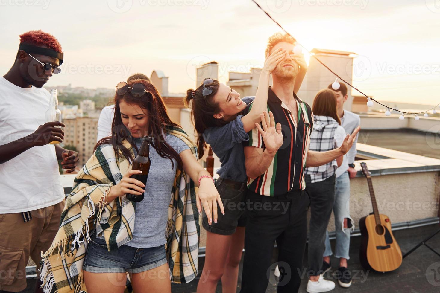 Playing game. Man's eyes covered by hands. Group of young people in casual clothes have a party at rooftop together at daytime photo
