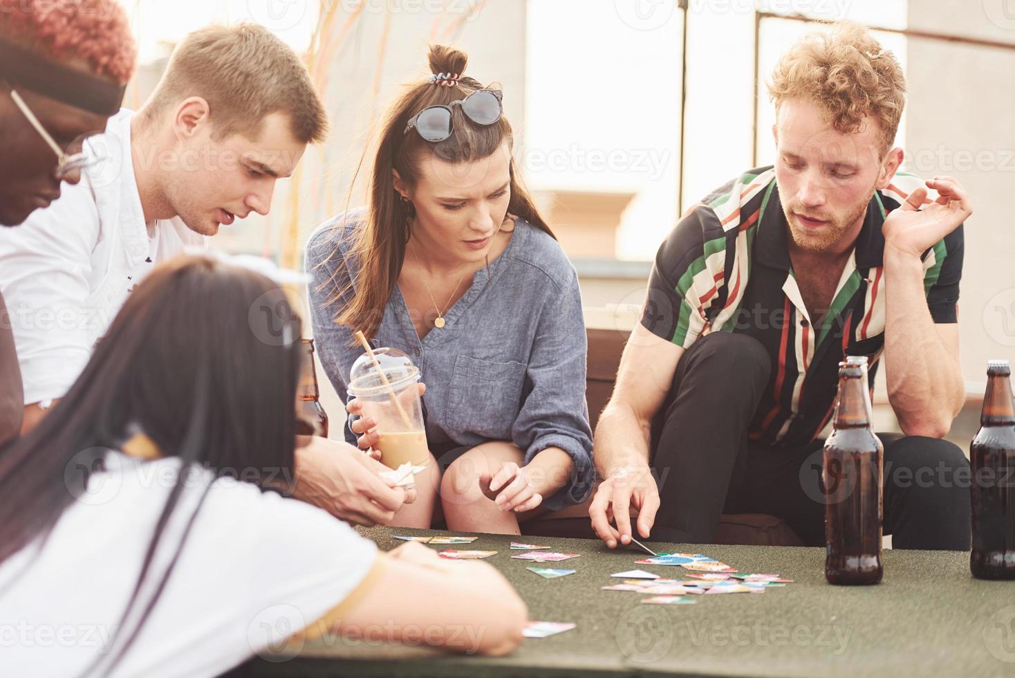 Playing game and sitting by the table. Group of young people in casual clothes have a party at rooftop together at daytime photo