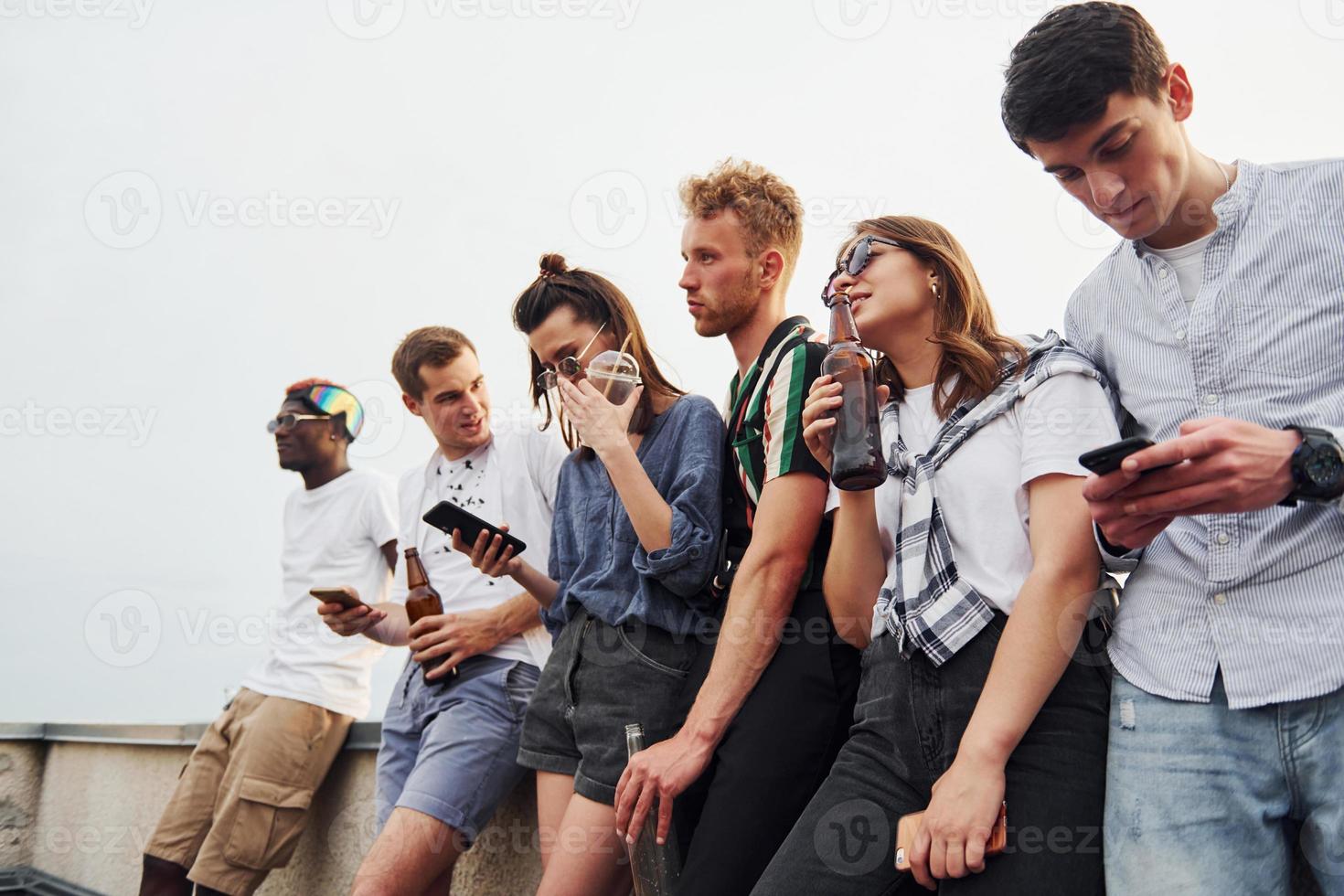 Standing with phones and alcohol in hands. Group of young people in casual clothes have a party at rooftop together at daytime photo