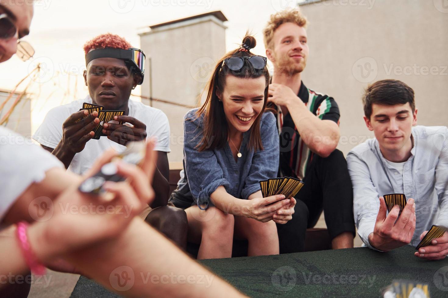 Sitting by table and playing card game. Group of young people in casual clothes have a party at rooftop together at daytime photo