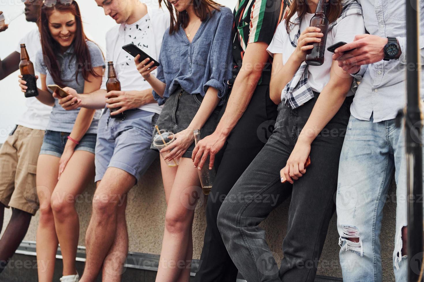 Standing with phones and alcohol in hands. Group of young people in casual clothes have a party at rooftop together at daytime photo