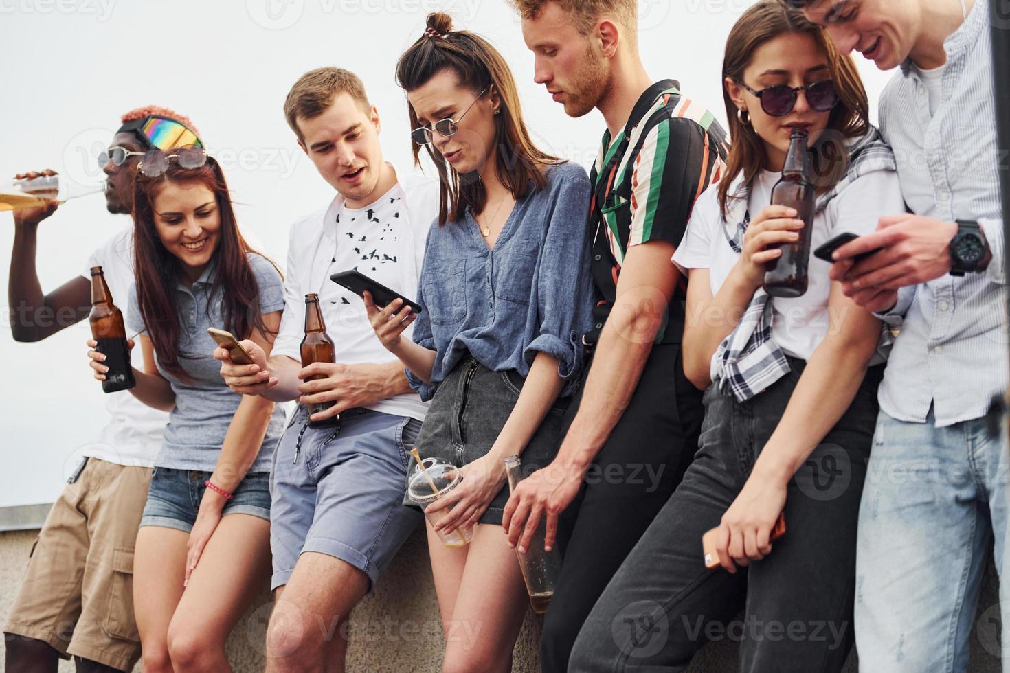 Standing with phones and alcohol in hands. Group of young people in casual clothes have a party at rooftop together at daytime photo