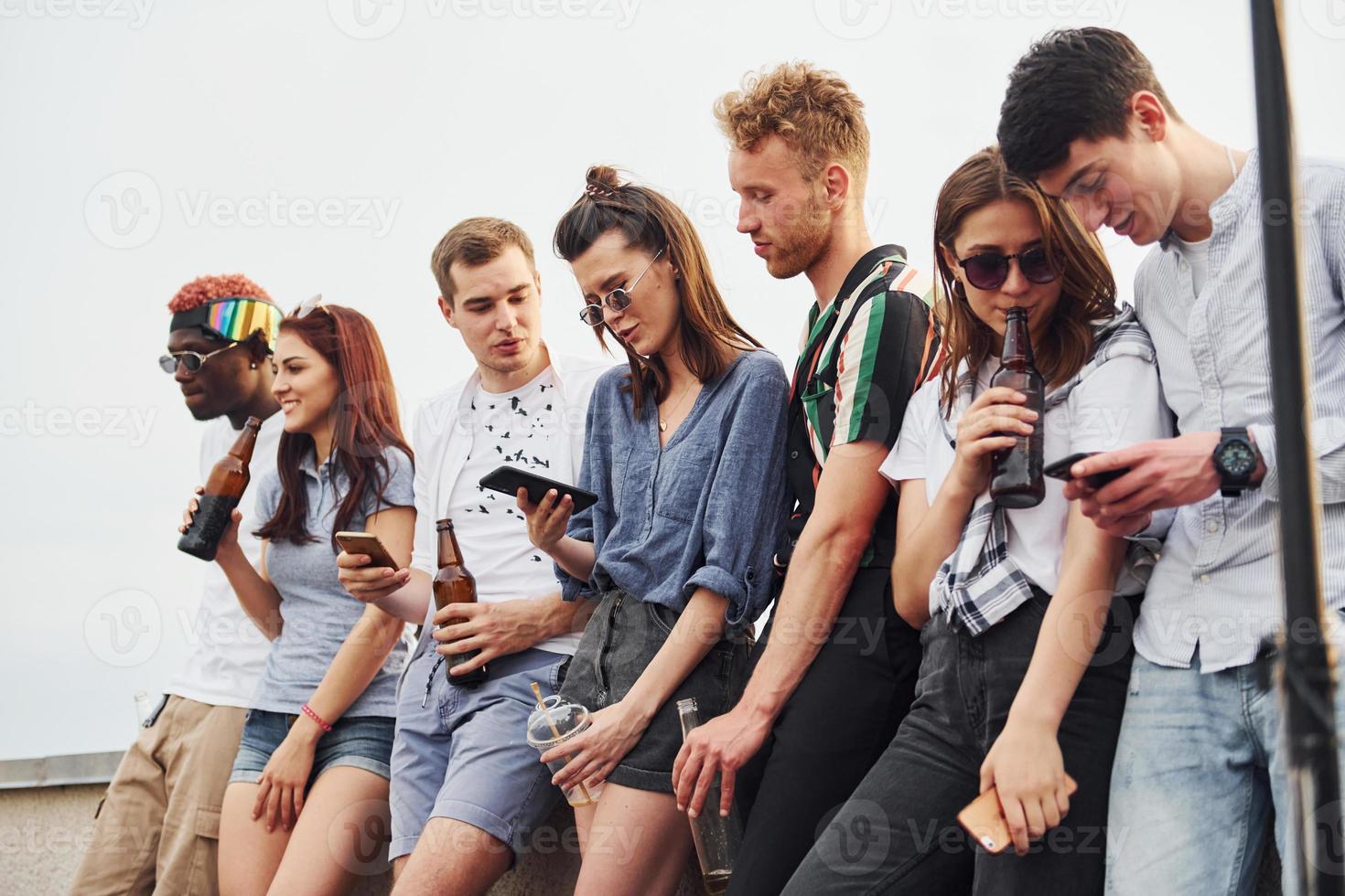 Standing with phones and alcohol in hands. Group of young people in casual clothes have a party at rooftop together at daytime photo