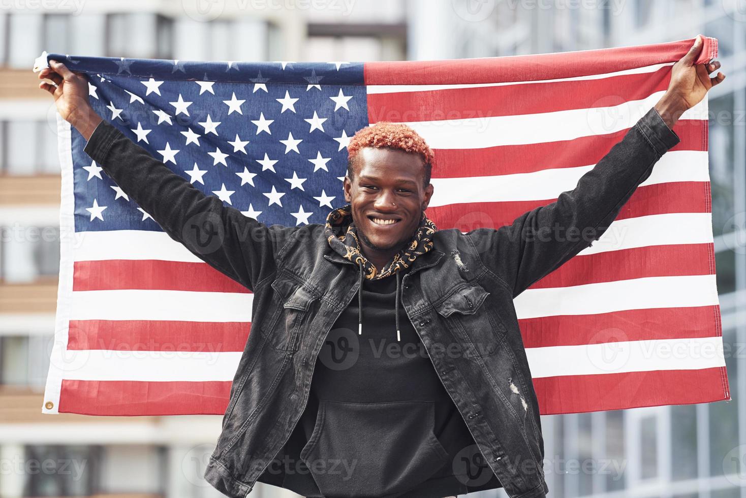 Patriot holding USA flag. Conception of pride and freedom. Young african american man in black jacket outdoors in the city standing against modern business building photo