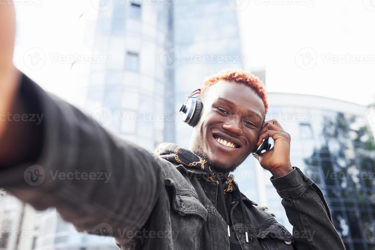 Listening to music in headphones. Young african american man in black jacket outdoors in the city standing against modern business building photo