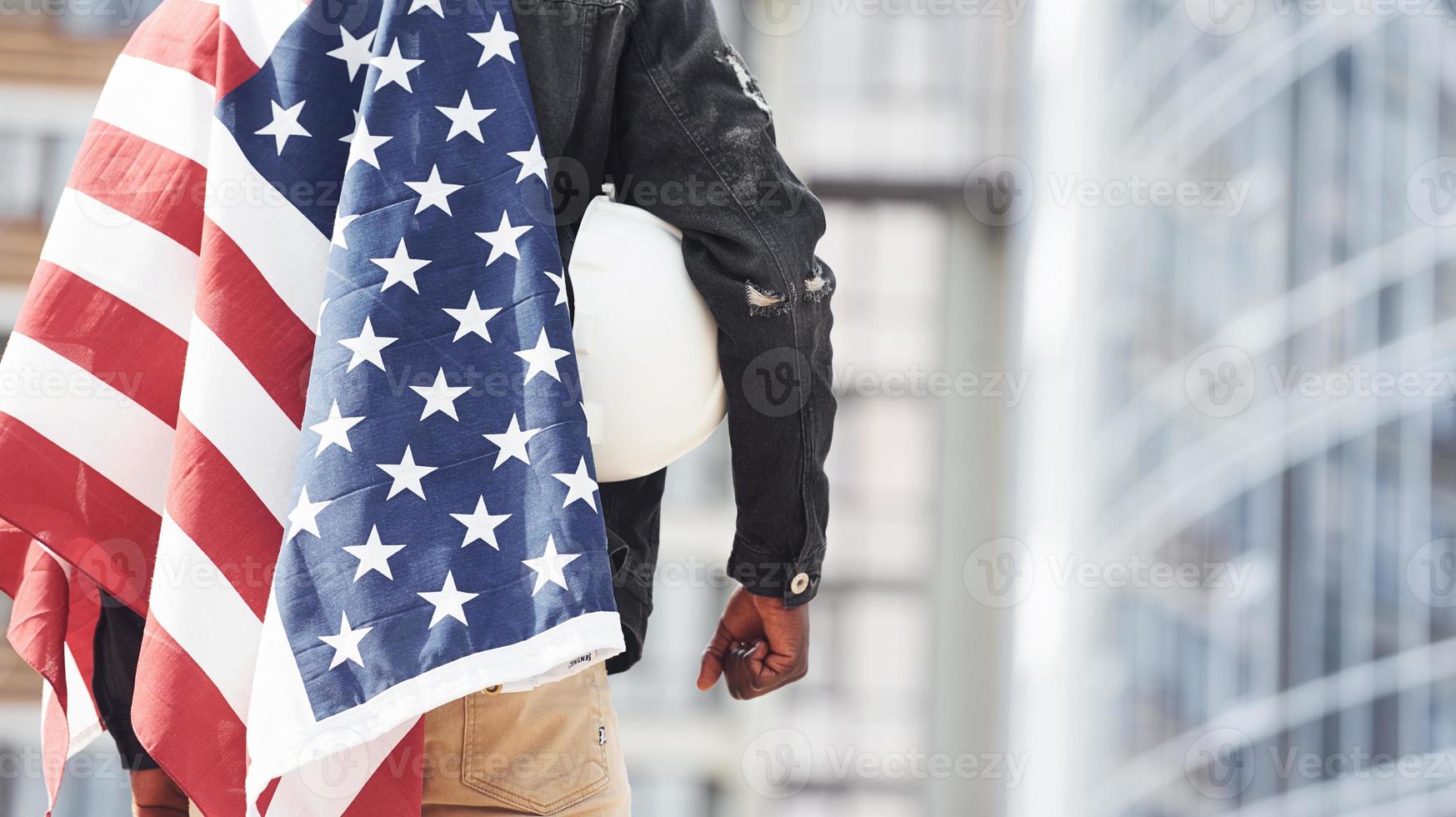 View from behind. Patriot holding USA flag. Conception of pride and freedom. Young african american man in black jacket outdoors in the city standing against modern business building photo