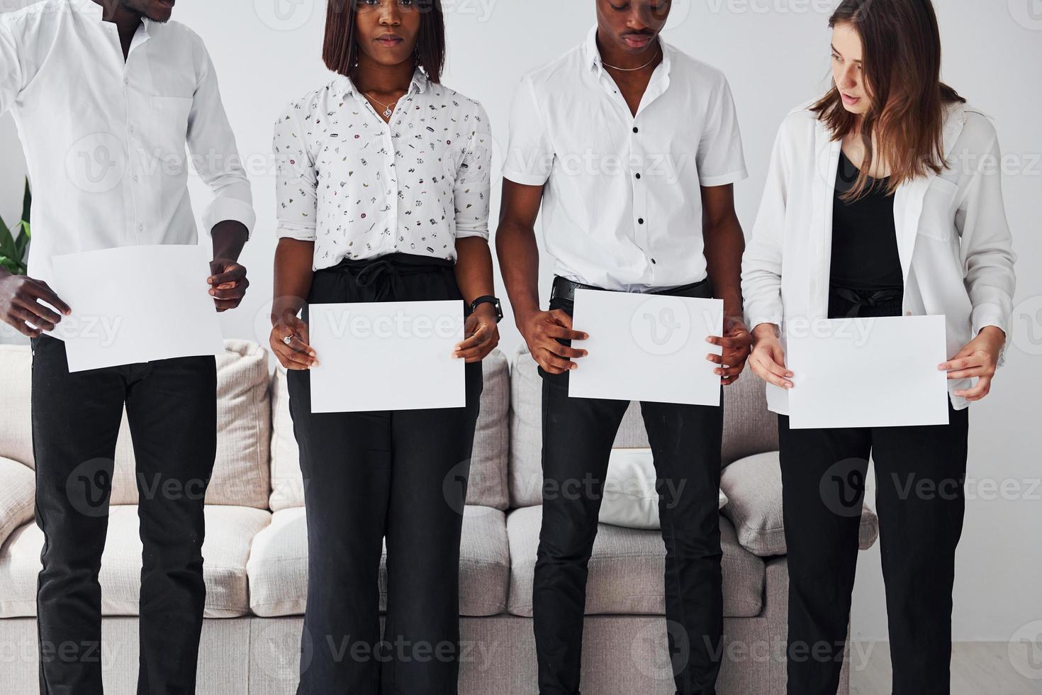 Group of african american people in formal clothes standing near sofa indoors in office together and holding empty papers in hands photo