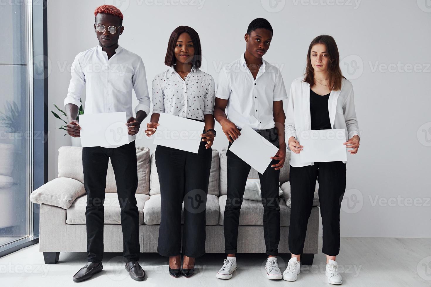 Group of african american people in formal clothes standing near sofa indoors in office together and holding empty papers in hands photo