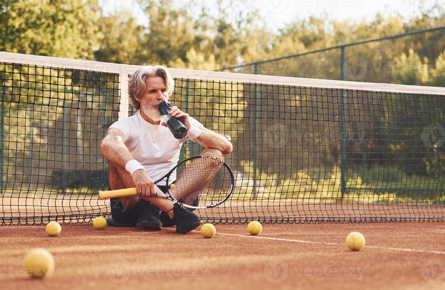 Taking break and drinking water. Senior stylish man in white shirt and black sportive shorts on tennis court photo
