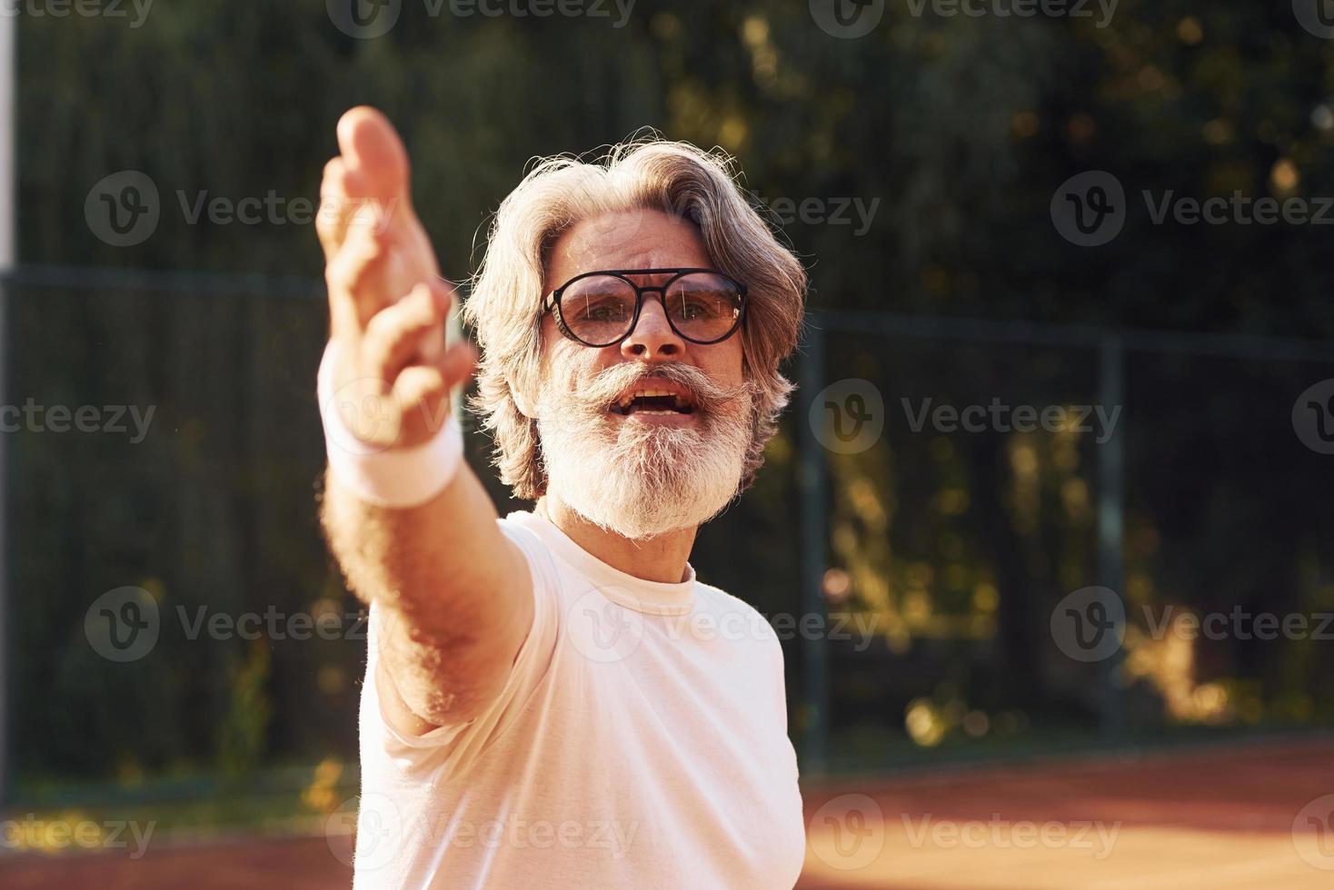 hombre mayor con estilo en anteojos, camisa blanca y pantalones cortos deportivos negros en la cancha de tenis foto