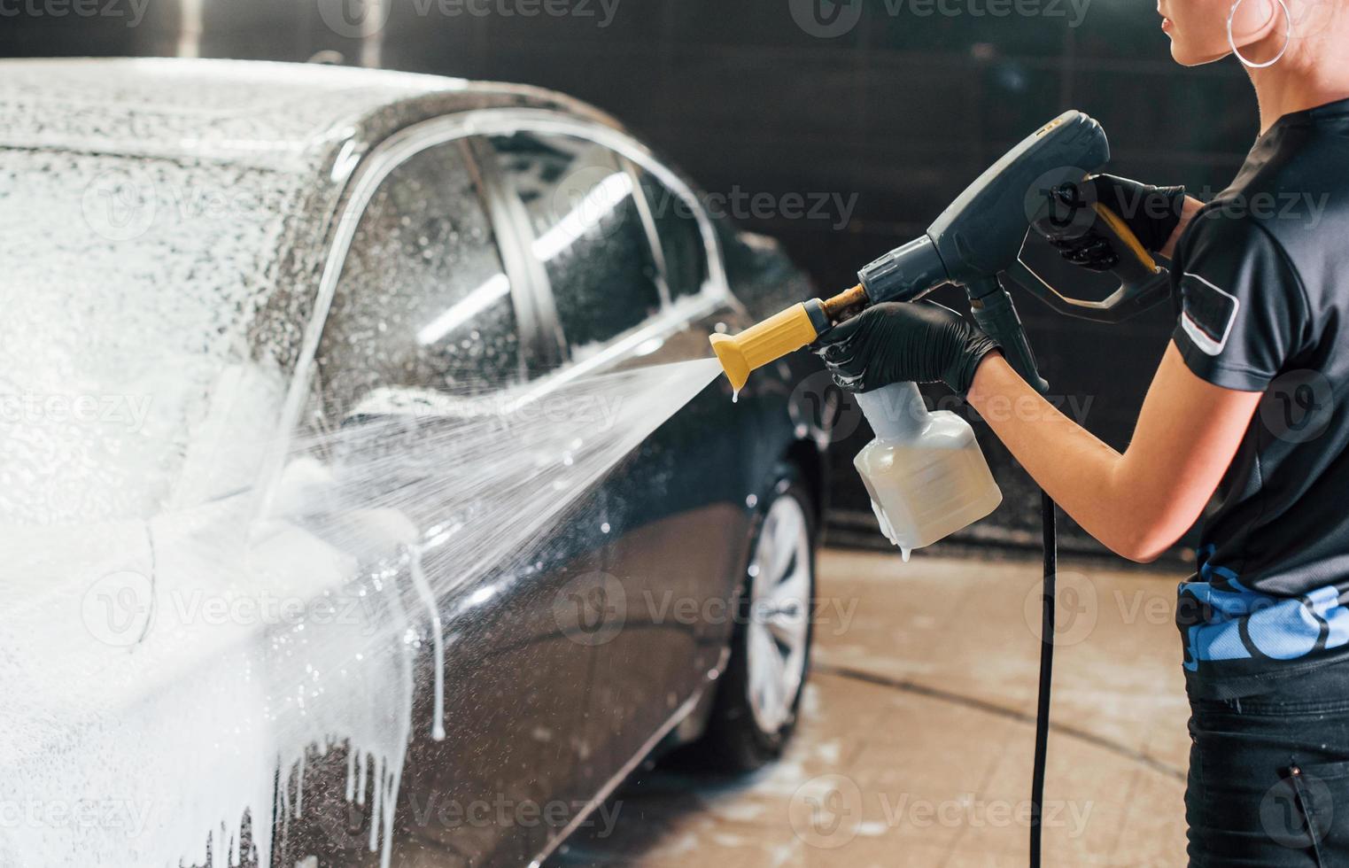 utilizando agua a alta presión. el automóvil negro moderno es limpiado por una mujer dentro de la estación de lavado de autos foto