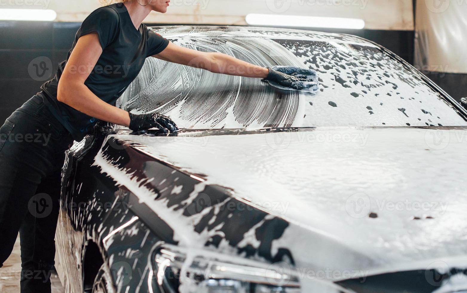 Wipes vehicle that is in white soap. Modern black automobile get cleaned by woman inside of car wash station photo