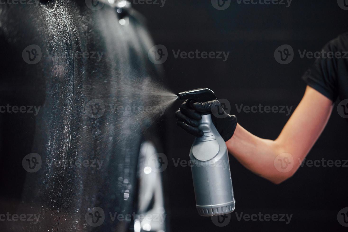 rociando el vehículo. el automóvil negro moderno es limpiado por una mujer dentro de la estación de lavado de autos foto