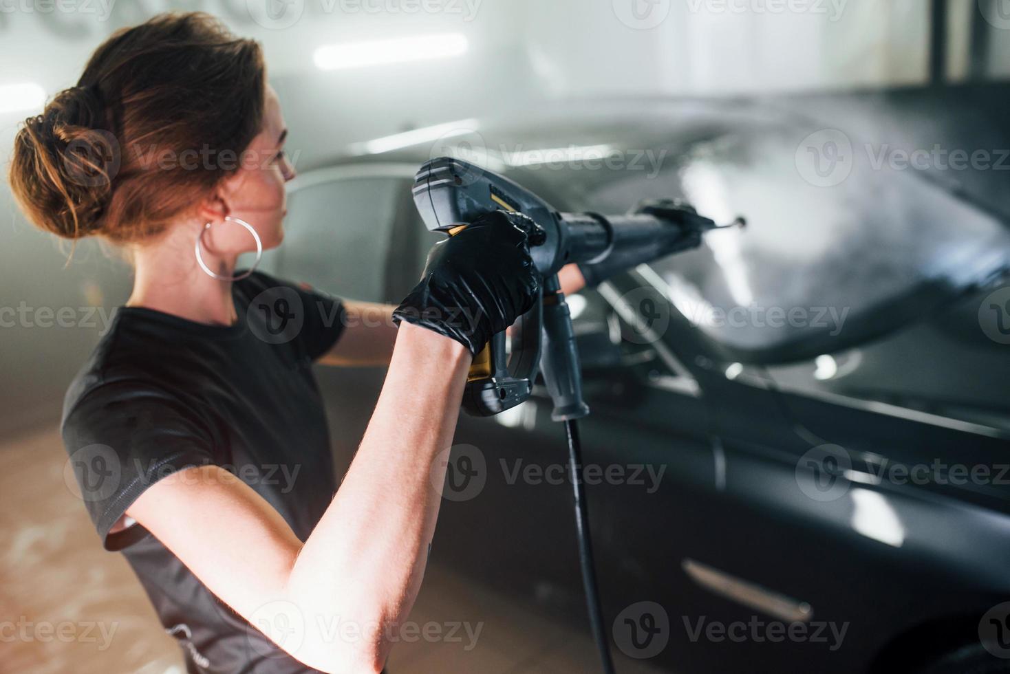 Using equipment with high pressure water. Modern black automobile get cleaned by woman inside of car wash station photo