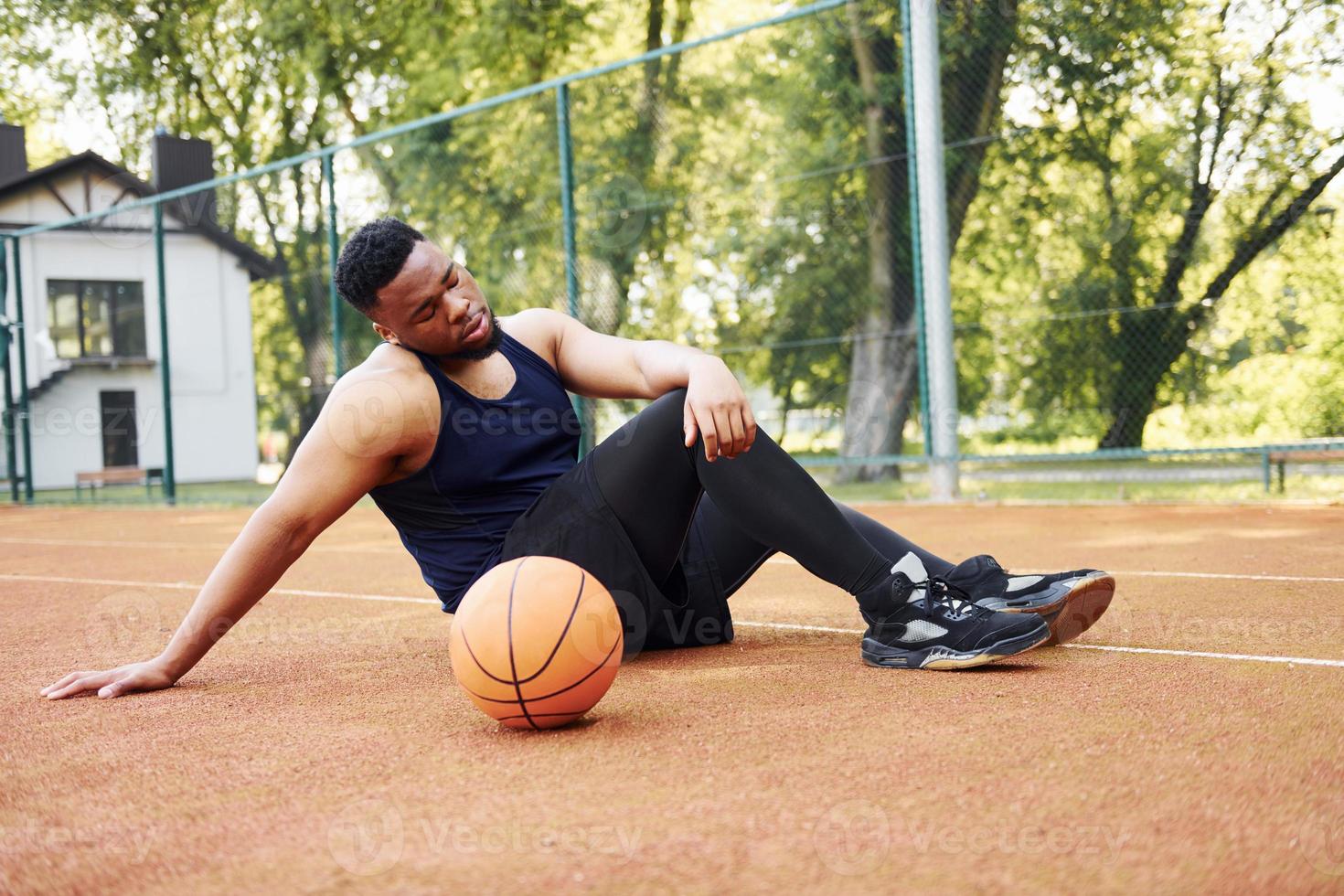 Tired african american man sits on the ground with ball on the court outdoors. Takes a break photo