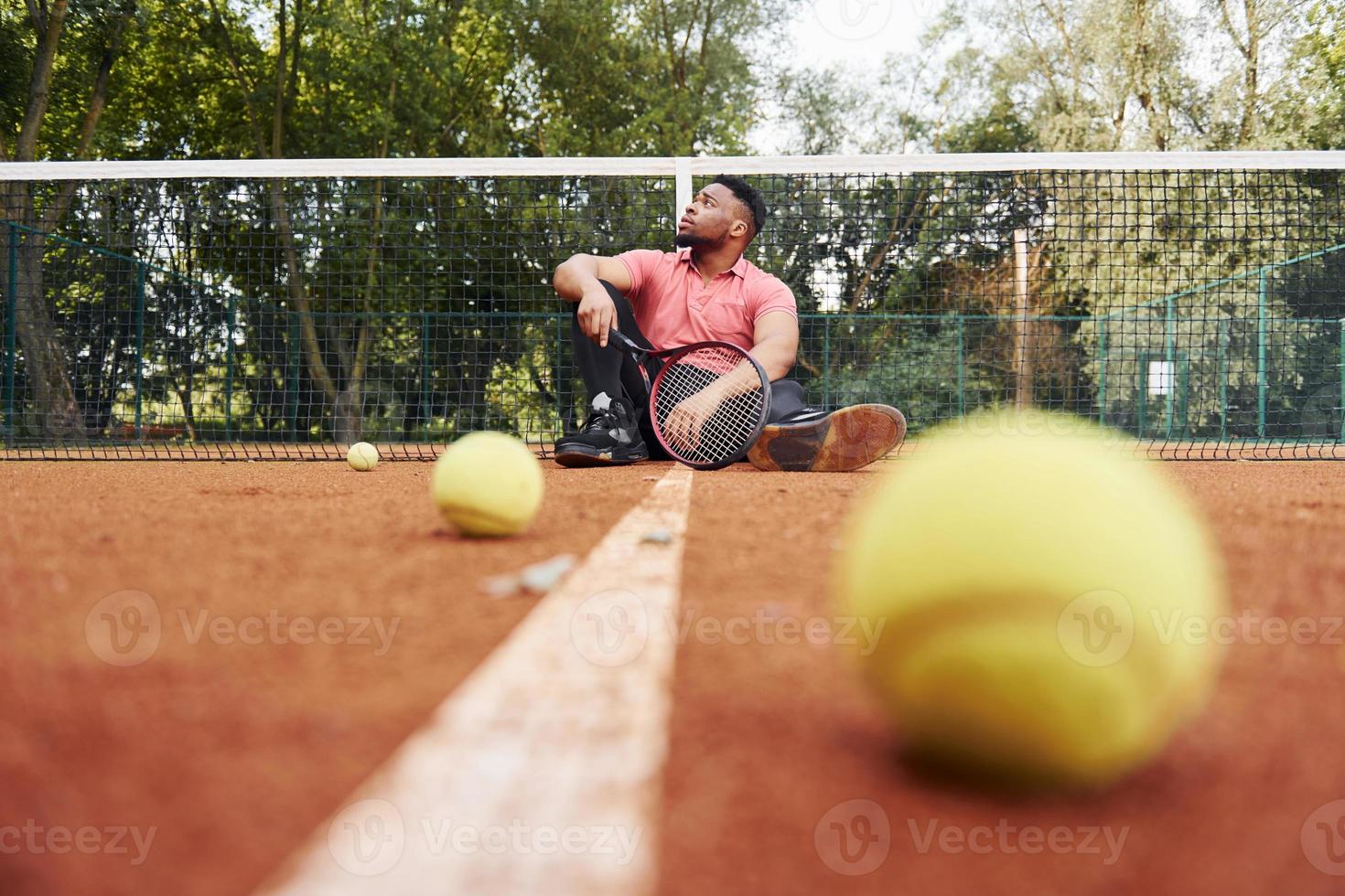 Sits near net and taking a break. African american man in pink shirt sits with tennis racket on the court outdoors photo