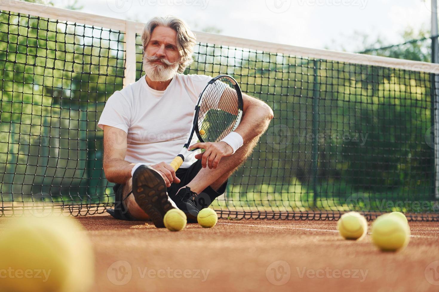 se sienta en el suelo y toma un descanso. Senior hombre moderno y elegante con raqueta al aire libre en la cancha de tenis durante el día foto