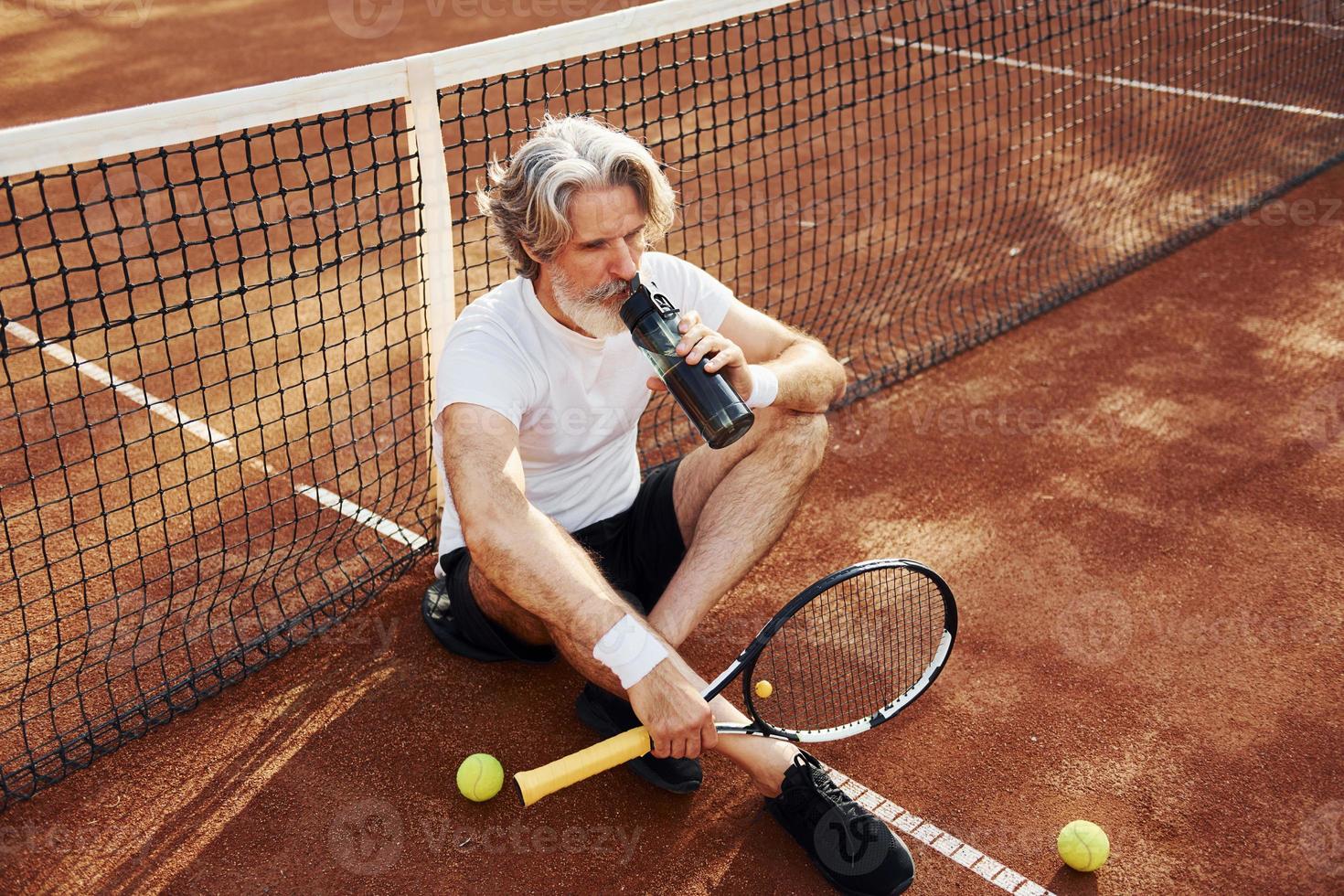 Drinking water and sitting on the ground. Senior modern stylish man with racket outdoors on tennis court at daytime photo