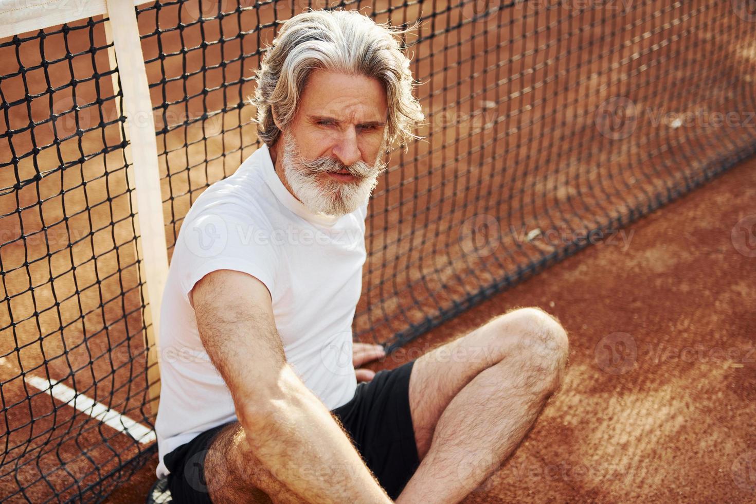 Sits on the ground and takes break. Senior modern stylish man with racket outdoors on tennis court at daytime photo