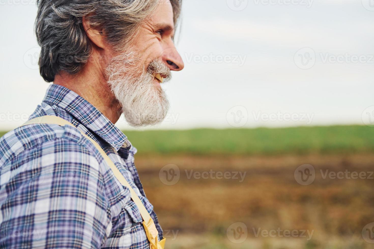 Side view. In yellow uniform. Senior stylish man with grey hair and beard on the agricultural field with harvest photo