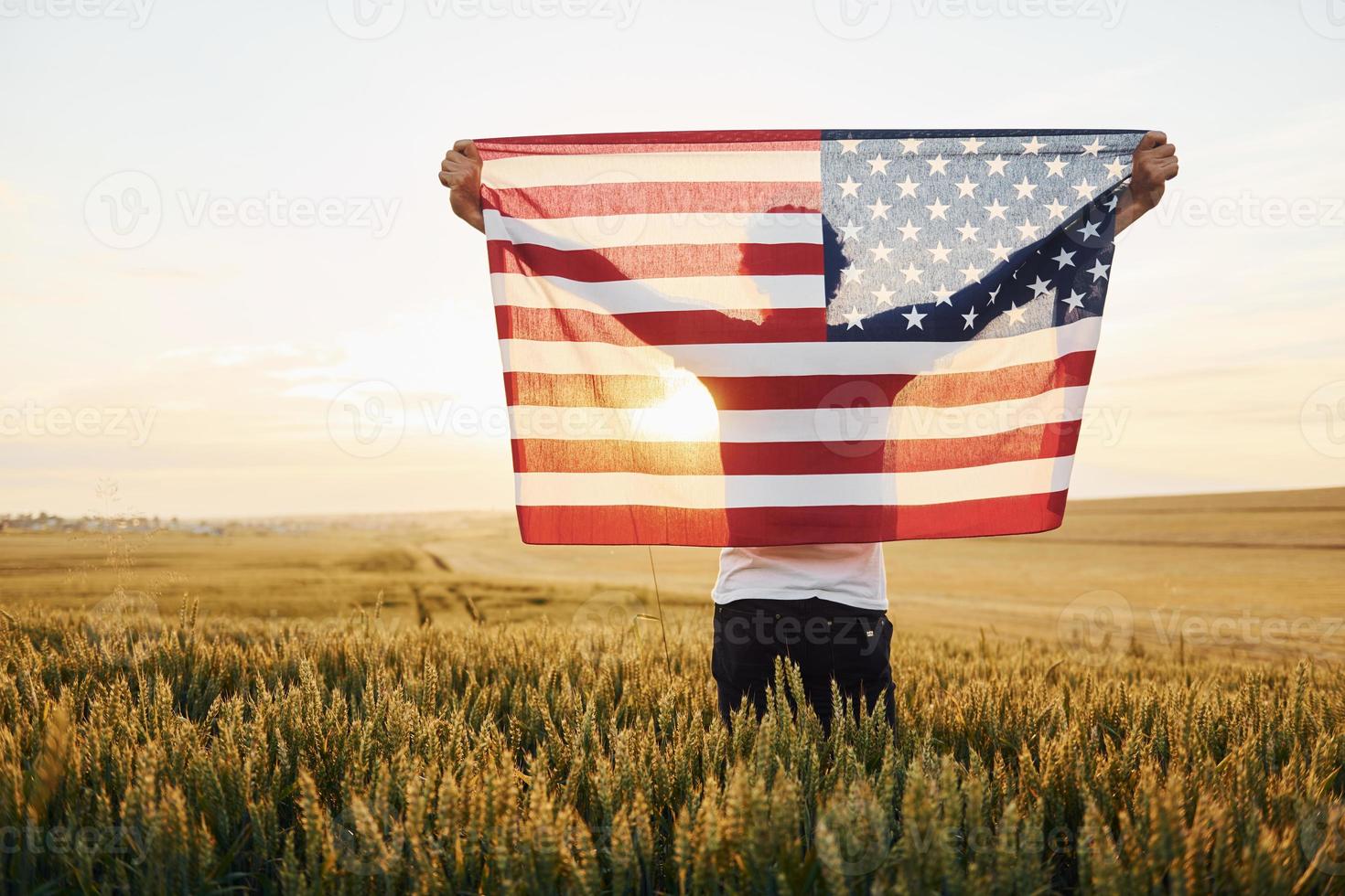 vista desde atrás. sosteniendo la bandera de estados unidos en las manos. hombre elegante patriótico senior con pelo gris y barba en el campo agrícola foto