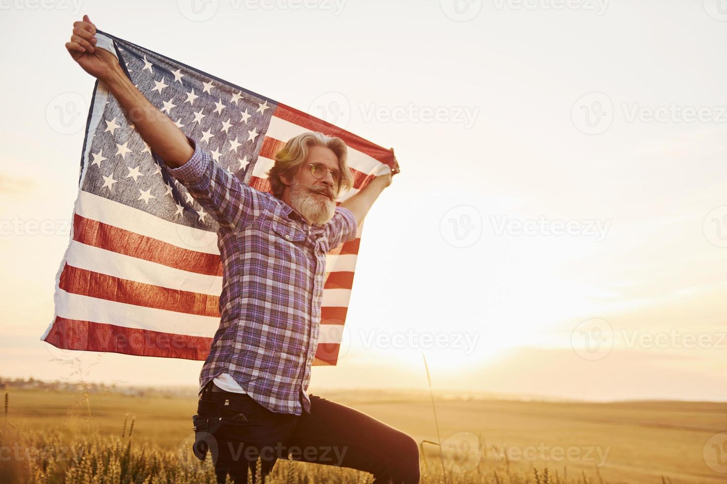 Holding USA flag in hands. Patriotic senior stylish man with grey hair and beard on the agricultural field photo