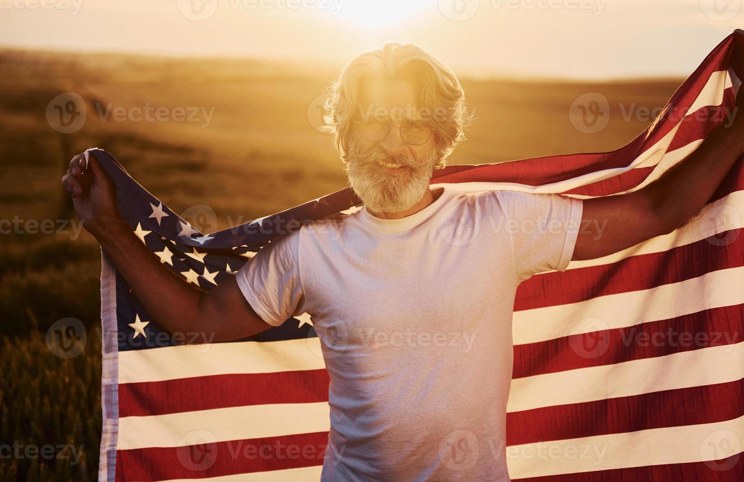 Conception of freedom. Holding USA flag. Senior stylish man with grey hair and beard on the agricultural field with harvest photo