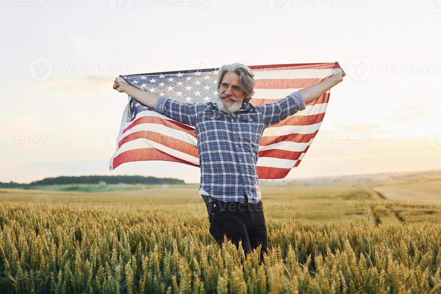 Holding USA flag in hands. Patriotic senior stylish man with grey hair and beard on the agricultural field photo