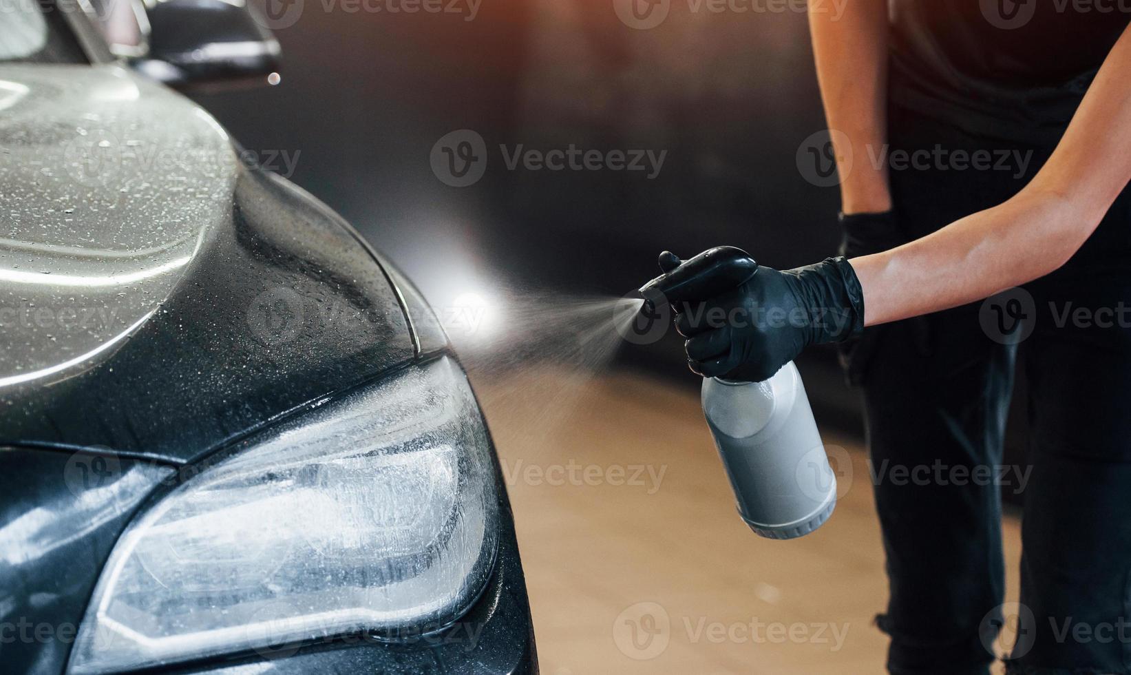 rociando el vehículo. el automóvil negro moderno es limpiado por una mujer dentro de la estación de lavado de autos foto