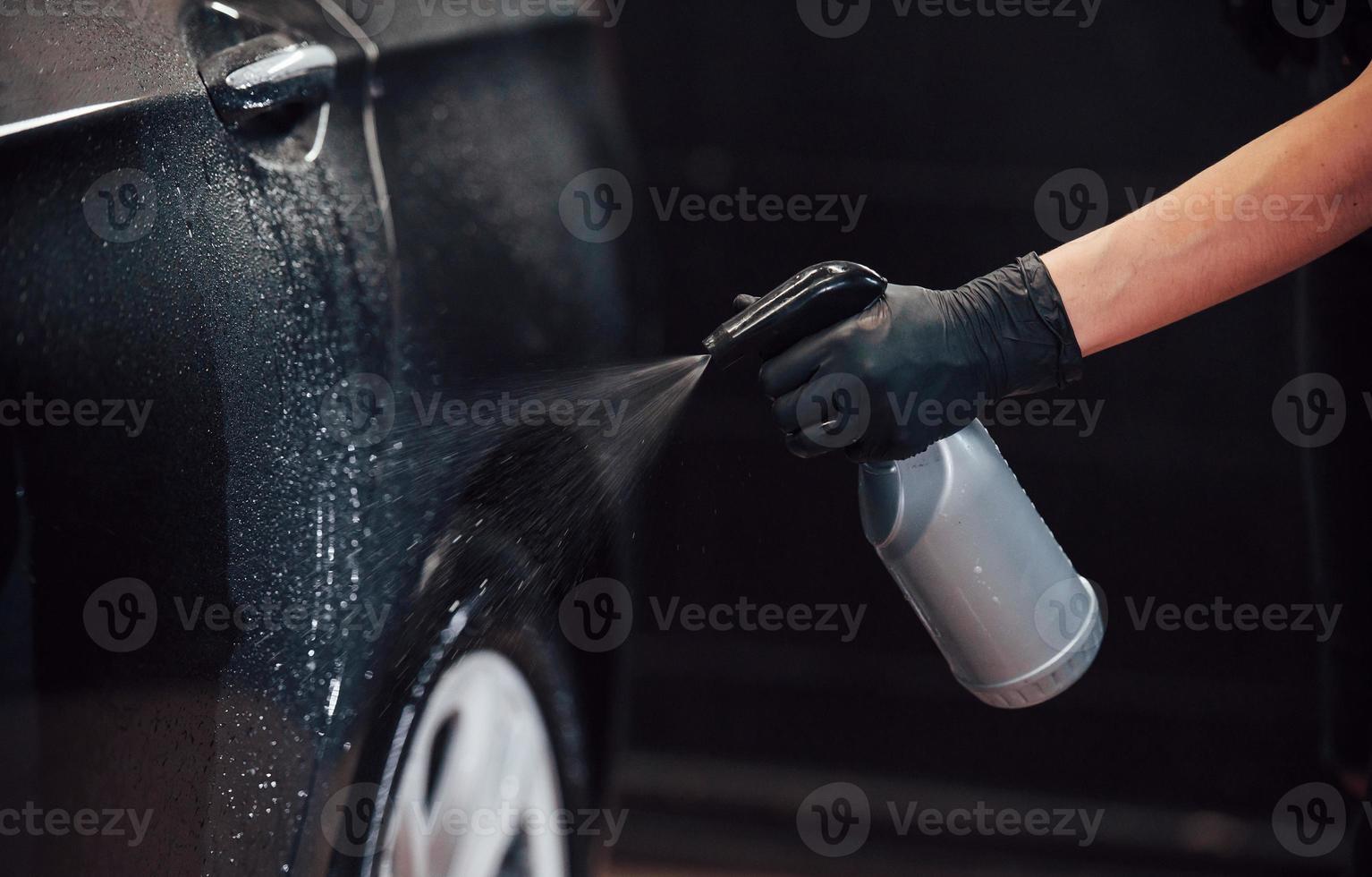 rociando el vehículo. el automóvil negro moderno es limpiado por una mujer dentro de la estación de lavado de autos foto